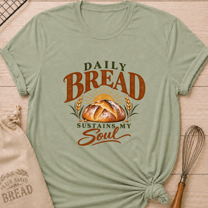 Sage women's relaxed fit tee with Daily Bread Sustains My Soul lettering around a golden artisan loaf beneath a rising sun, framed by wheat in rich earthy tones