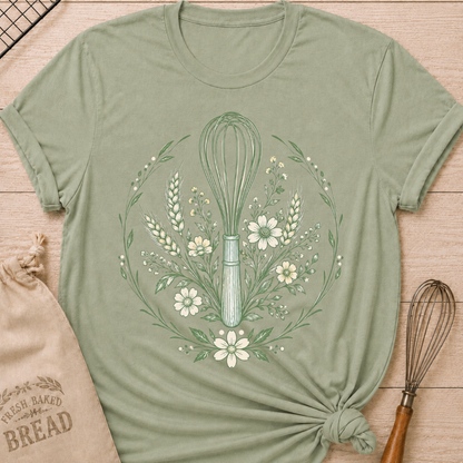 A sage graphic t-shirt styled with a knotted hem, laid flat on a light wood background with a drawstring bread bag and a whisk. The shirt features a detailed botanical illustration of a kitchen whisk centered within a circular wreath of wildflowers, wheat stalks, daisies, and delicate foliage, all rendered in sage green line art.