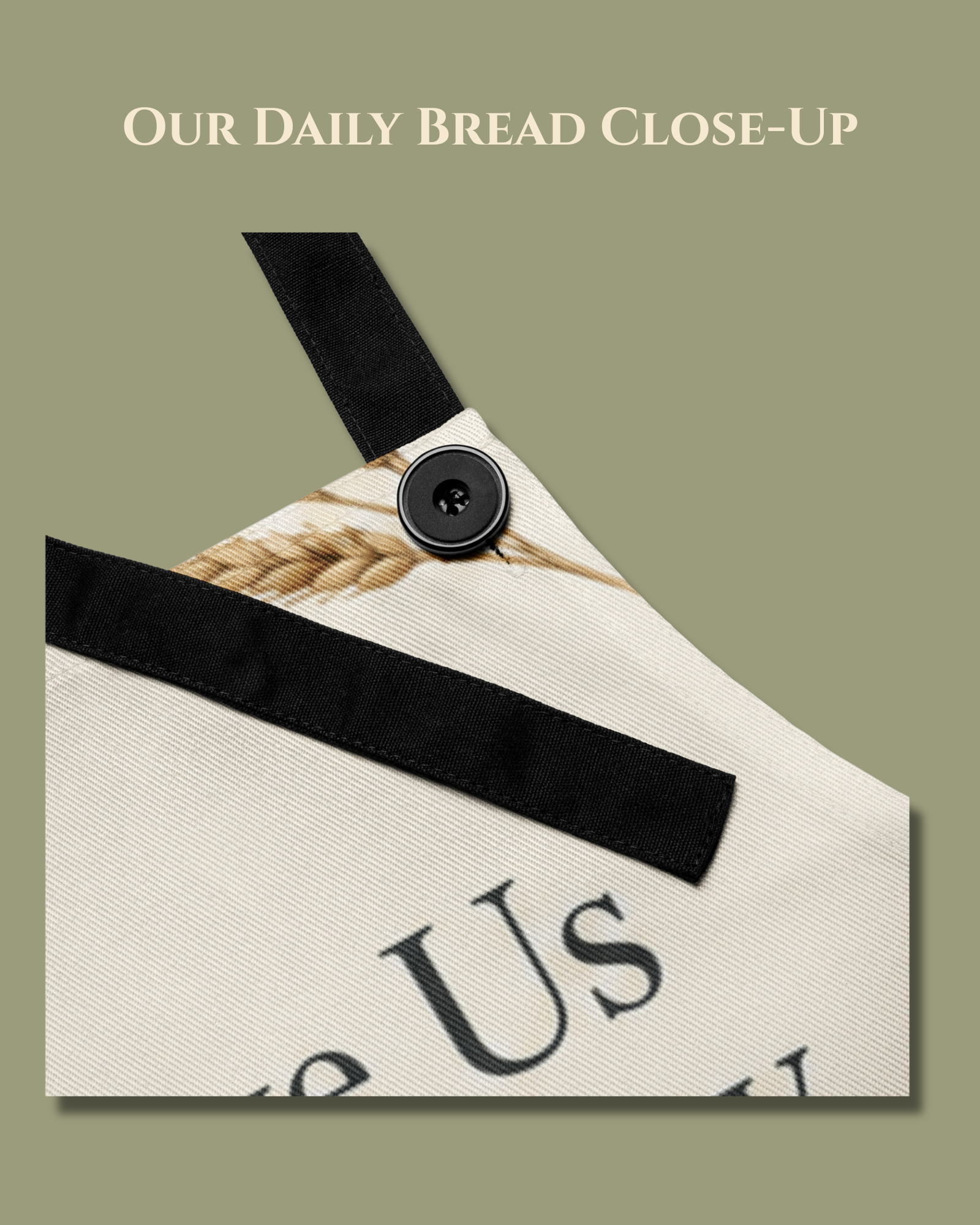Close-up of the Our Daily Bread apron showing the black adjustable neck strap and button detail against the cream fabric with its wheat illustration.