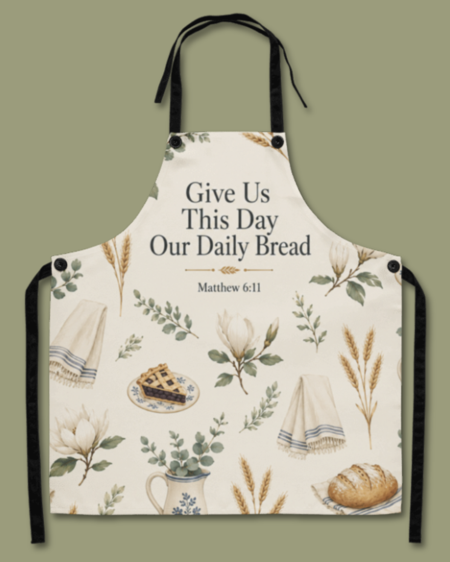 An apron with a warm cream background printed with botanical illustrations of wheat, flowers, a pie, a teacup, and a loaf of bread. The center features the text 'Give Us This Day Our Daily Bread' and 'Matthew 6:11'.
