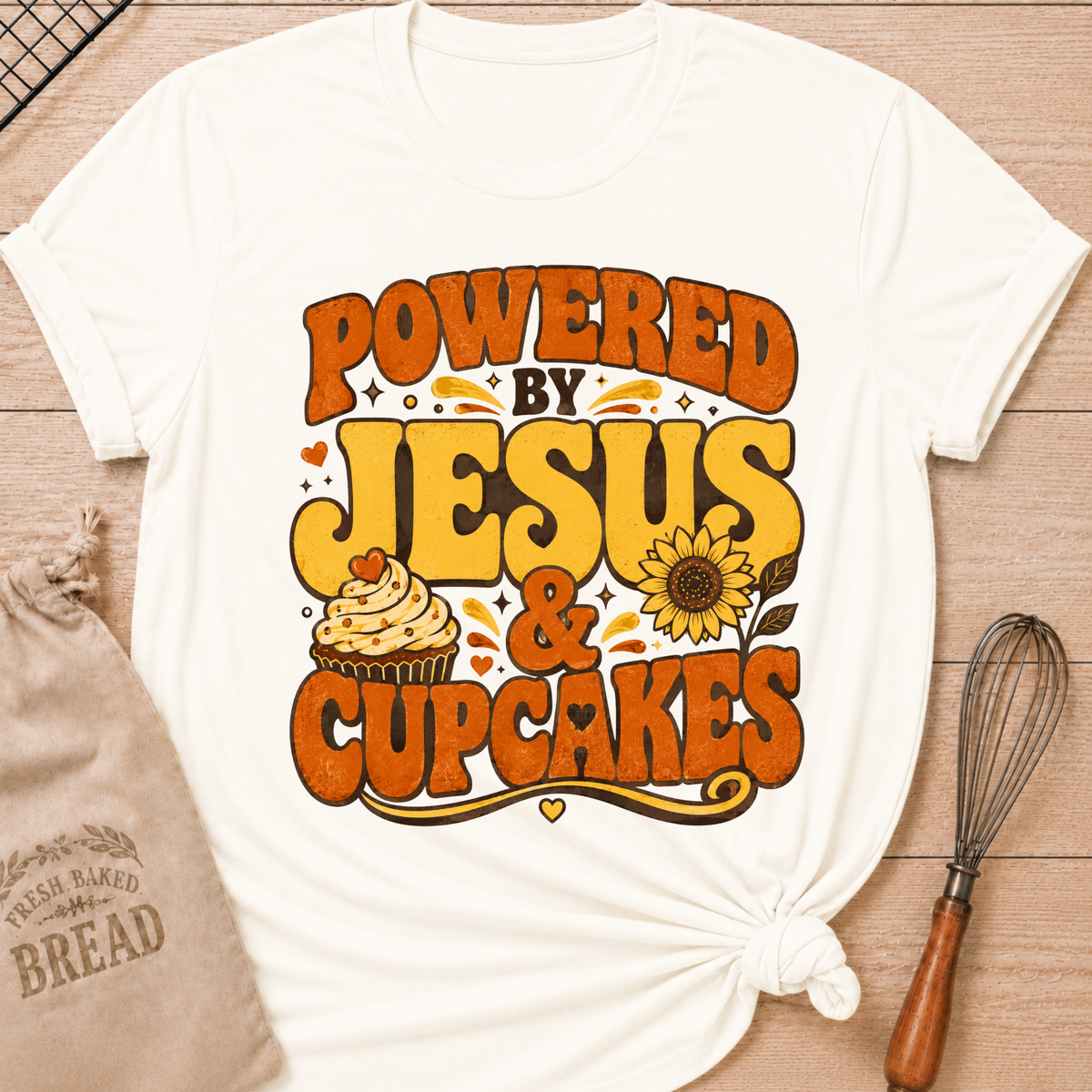A white graphic t-shirt with a retro-style orange and yellow print reading 'POWERED BY JESUS & CUPCAKES' with a cupcake and sunflower illustration, styled flat-lay on a wooden surface with a bread bag and whisk nearby.