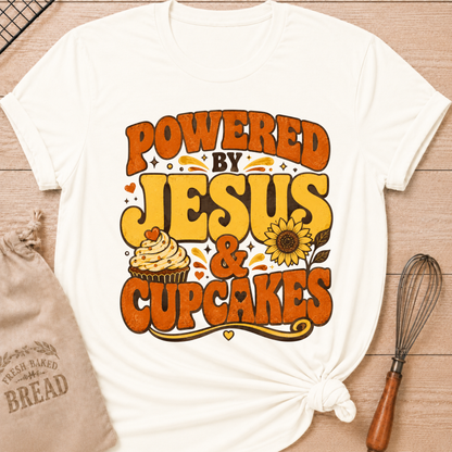 A white graphic t-shirt with a retro-style orange and yellow print reading 'POWERED BY JESUS & CUPCAKES' with a cupcake and sunflower illustration, styled flat-lay on a wooden surface with a bread bag and whisk nearby.