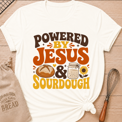 An off-white graphic t-shirt with a 'POWERED BY JESUS & SOURDOUGH' design, styled in a flat-lay on a wooden background with a burlap bag, bread, and a whisk. graphic t-shirt laid flat on a light wood background, styled with a bread bag, whisk, and baking paper. The t-shirt features a graphic that reads 'POWERED BY JESUS & SOURDOUGH' in brown and orange lettering, accompanied by illustrations of a sourdough bread loaf, a mason jar of dough, and a sunflower.