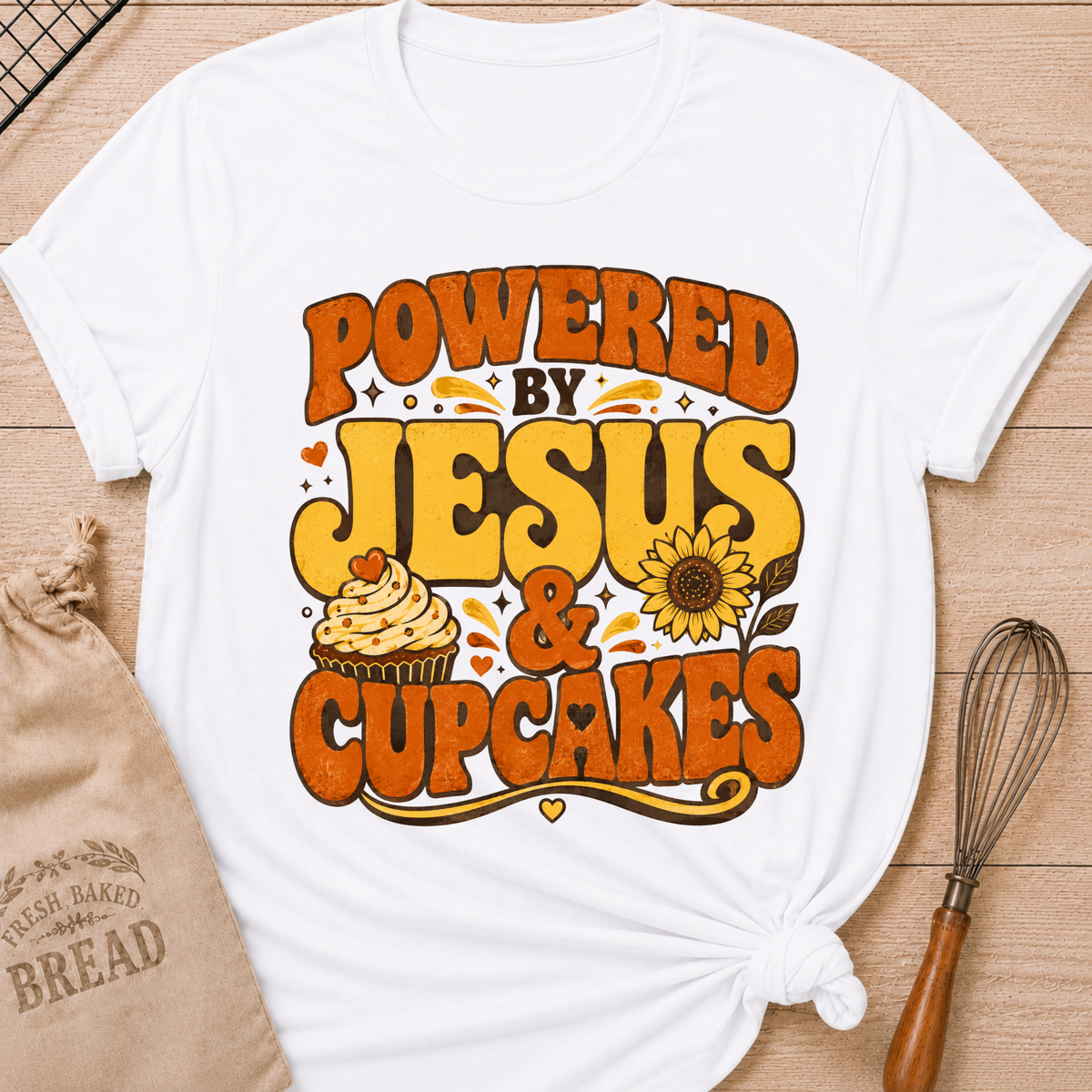 A white short-sleeve T-shirt laid flat on a wood surface, styled with a kraft bread bag, a whisk, and a printed graphic that reads “POWERED BY JESUS & CUPCAKES” in orange and yellow lettering with a cupcake and sunflower illustration.