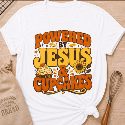 A white short-sleeve T-shirt laid flat on a wood surface, styled with a kraft bread bag, a whisk, and a printed graphic that reads “POWERED BY JESUS & CUPCAKES” in orange and yellow lettering with a cupcake and sunflower illustration.