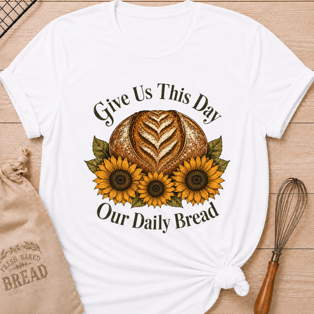 Women's relaxed fit bright white t-shirt laid flat on a light wood surface, featuring a faith-inspired graphic of a rustic sourdough boule with a heart-shaped wheat score surrounded by three golden sunflowers and deep olive green leaves, with the text "Give Us This Day Our Daily Bread" in a classic serif font. Styled alongside a linen "Fresh Baked Bread" drawstring bag and a vintage-handled wire whisk.