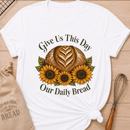 Women's relaxed fit bright white t-shirt laid flat on a light wood surface, featuring a faith-inspired graphic of a rustic sourdough boule with a heart-shaped wheat score surrounded by three golden sunflowers and deep olive green leaves, with the text "Give Us This Day Our Daily Bread" in a classic serif font. Styled alongside a linen "Fresh Baked Bread" drawstring bag and a vintage-handled wire whisk.