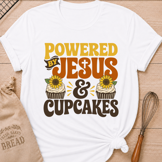 A white short-sleeve T-shirt laid flat on a wood surface, styled with a knotted hem, a burlap bread bag, and a whisk. The shirt features a large decorative graphic reading “POWERED BY JESUS & CUPCAKES” in yellow, orange, brown, and black, with two cupcake illustrations and sunflower motifs.