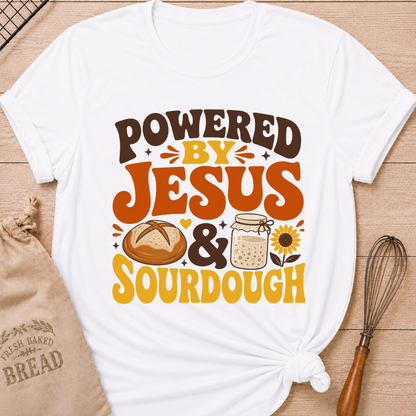 A white graphic t-shirt with a 'POWERED BY JESUS & SOURDOUGH' design, styled in a flat-lay on a wooden background with a burlap bread bag, a whisk, and a small sunflower illustration.