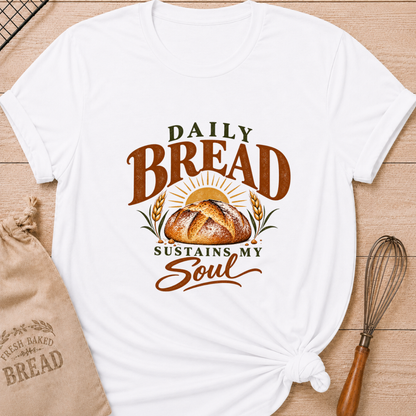 White women's relaxed fit tee with Daily Bread Sustains My Soul lettering around a golden artisan loaf beneath a rising sun, framed by wheat in rich earthy tones